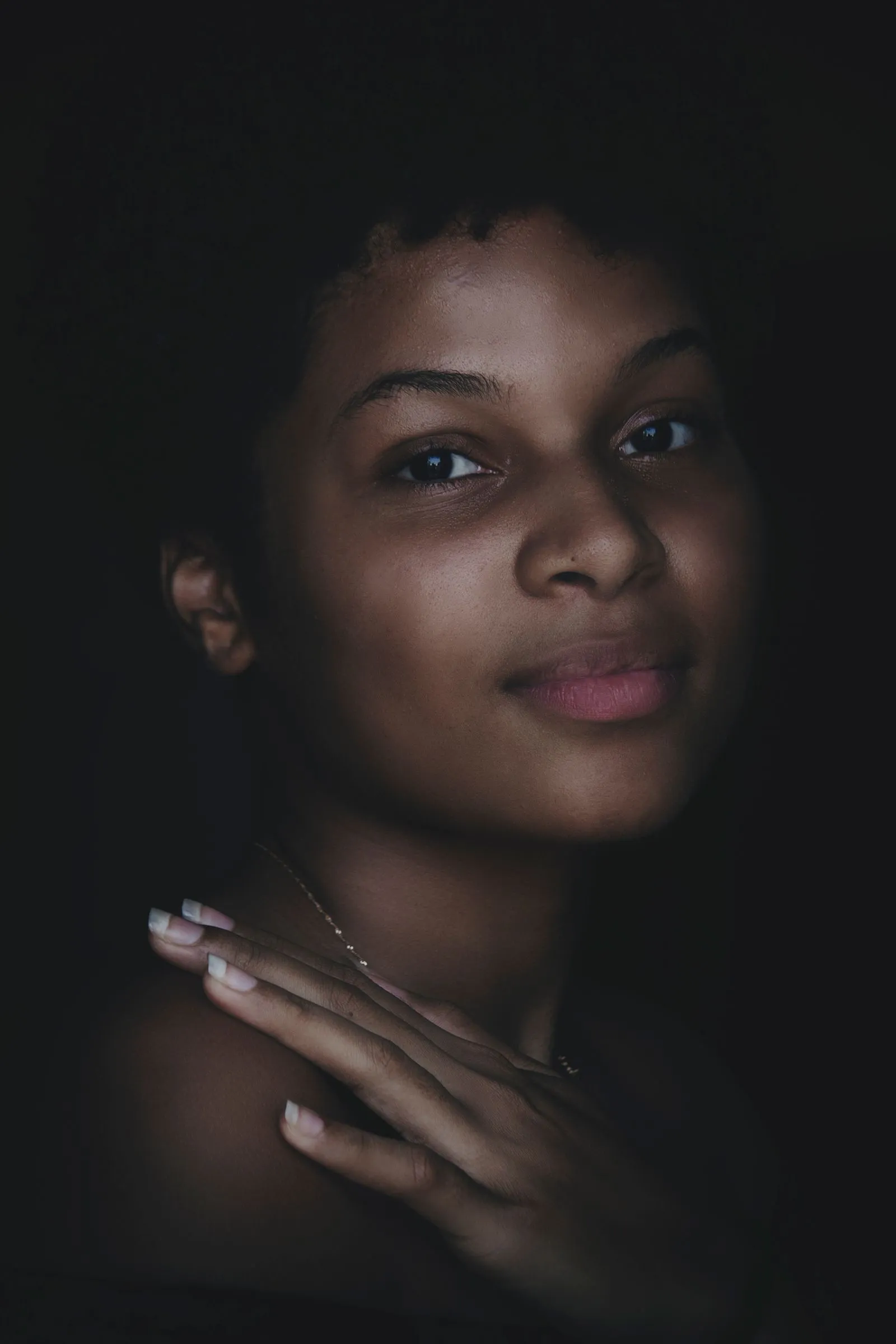 A young Black woman with a soft, hopeful expression and her hand resting on her shoulder, lit gently against a calm, dark background